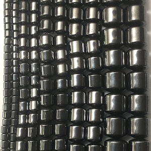 May include: Rows of dark gray, cylindrical beads with a metallic sheen. The beads are arranged in vertical lines, varying in size from small to large. The beads are likely for jewelry making.