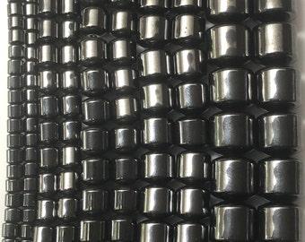 May include: Rows of dark gray, cylindrical beads with a metallic sheen. The beads are arranged in vertical lines, varying in size from small to large. The beads are likely for jewelry making.