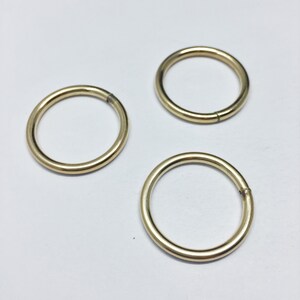 May include: Three gold-colored metal rings are displayed against a white background. The rings are circular, with a smooth, shiny surface. Each ring has a small gap, indicating a possible opening or closure mechanism. The rings appear to be new.