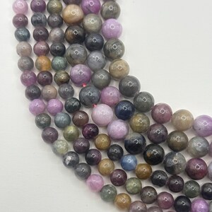May include: Strands of multi-colored gemstone beads. The beads are round and vary in size, with colors including pink, purple, brown, green, and gray. The beads are strung together, creating a textured and colorful display. Ideal for jewelry making.
