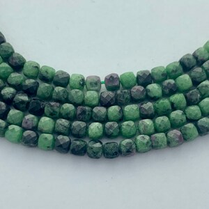 May include: Close-up of strands of faceted beads. The beads are a mix of green and dark green with flecks of red. The beads are square-shaped and strung together with green thread. The background is white.