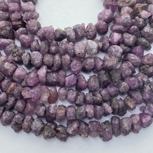 May include: Strands of rough-cut, tumbled amethyst gemstone beads. The beads are a mix of light and dark purple hues, with a natural, irregular shape. The beads are strung together on a red string, ready for jewelry making or other crafts.