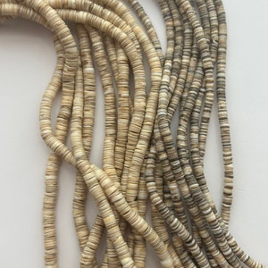 May include: Strands of natural, flat, disc-shaped beads in varying shades of cream, tan, and gray. The beads are strung together, creating a flowing, organic texture. Ideal for jewelry making or craft projects.