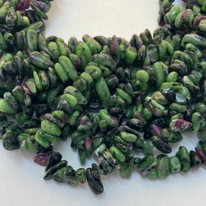 May include: Close-up of a collection of tumbled gemstones. The stones are a mix of green and dark green with flecks of red and purple. The gemstones are irregularly shaped and appear to be strung together.