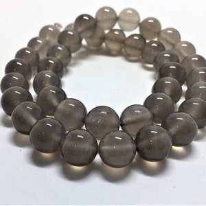 May include: A strand of round, smoky quartz beads. The beads are a translucent gray color and appear to be polished. The beads are strung together, forming a circular shape. The beads are approximately 10mm in diameter.