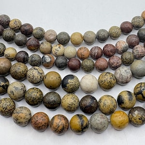 May include: Close-up of a collection of round, natural stone beads in various earth tones. The beads have a speckled pattern with colors ranging from beige and tan to brown and gray. The beads are strung together, suggesting they are for jewelry making.