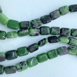 May include: Strands of square-shaped beads in shades of green and dark red. The beads have a polished, smooth surface and are strung together, creating a pattern. The beads are likely used for jewelry making.