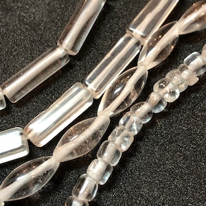 May include: Close-up of several strands of clear glass beads. The beads vary in shape, including elongated cylinders, ovals, and small square beads. Each bead is strung on a white cord, creating a delicate and transparent appearance.