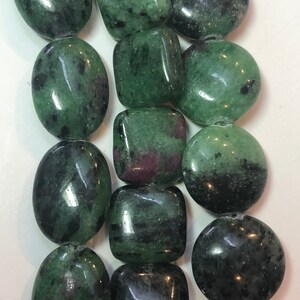 May include: Close-up of a strand of polished gemstone beads. The beads are a mix of oval, square, and round shapes. The stones are a deep green with black and red inclusions, giving them a unique, natural appearance.
