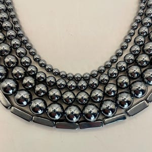 May include: Close-up of several strands of dark gray hematite beads. The beads vary in size, with the largest at the bottom and the smallest at the top. The beads have a shiny, reflective surface, and the strands are arranged in a curved, layered pattern.