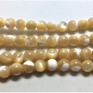 May include: Strands of round, iridescent beads in shades of cream and pale yellow. The beads have a glossy finish and a mix of solid and pearlescent surfaces. These beads are suitable for jewelry making.