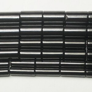 May include: Close-up of a collection of black, cylindrical beads. The beads are arranged in neat rows, each bead reflecting light. The beads appear to be made of a smooth, polished material. The overall effect is a dark, glossy texture.