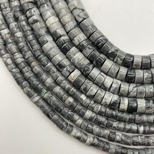May include: Close-up of strands of gray and black cylindrical beads. The beads have a marbled appearance with variations in color, ranging from light gray to deep charcoal. The beads are strung together, creating a textured and layered effect.