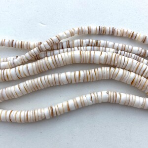 May include: Strands of white and tan shell beads. The beads are flat, disc-shaped, and strung together, creating a wavy pattern. The beads are approximately 0.25 inches in diameter, with a natural, organic appearance.