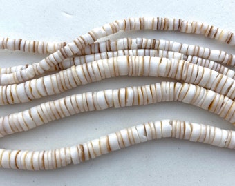 May include: Strands of white and tan shell beads. The beads are flat, disc-shaped, and strung together, creating a wavy pattern. The beads are approximately 0.25 inches in diameter, with a natural, organic appearance.