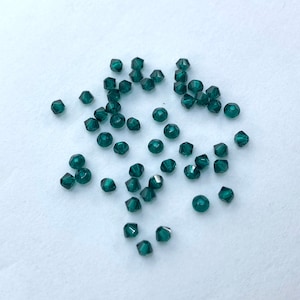 May include: A scattering of faceted, dark teal beads. The beads have a multifaceted design, reflecting light. The beads are small and round, with a polished, shiny appearance. They are scattered on a white surface. Ideal for jewelry making.