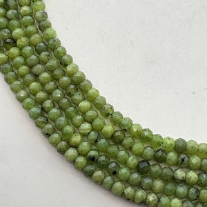 May include: Close-up of several strands of faceted, round, olive-green gemstone beads. The beads have a slightly speckled appearance and are strung together, creating a textured and organic aesthetic. The beads are a light green color.