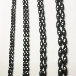 Peut inclure: Quatre rang&eacute;es de perles sombres et brillantes, de forme ovale, sont pr&eacute;sent&eacute;es sur fond blanc. Les perles sont enfil&eacute;es sur un fil rouge, avec des arrangements vari&eacute;s. Les perles semblent &ecirc;tre faites d'un mat&eacute;riau lisse et r&eacute;fl&eacute;chissant.