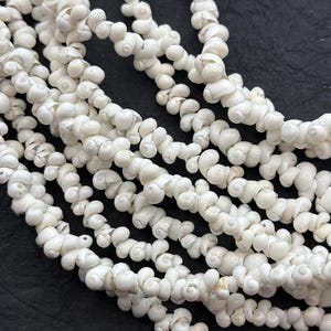 May include: Close-up of multiple strands of white seashell beads. The beads are small, spiral-shaped, and strung together, creating a textured, organic look. The background is a dark, solid color, which contrasts with the bright white beads.