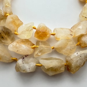 May include: Close-up of a necklace featuring rough-cut, light yellow citrine gemstones. The stones are strung together with gold-colored thread, creating a natural and earthy aesthetic. The citrine stones vary in size and shape.