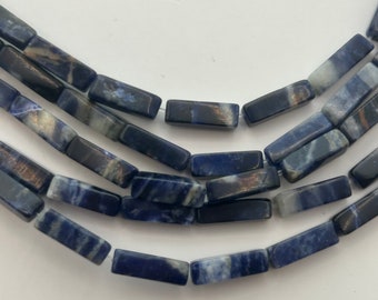 May include: Close-up of several strands of rectangular sodalite beads. The beads are a mix of dark blue, gray, and white, with a polished finish. The beads are strung together, creating a layered effect. The background is plain white.