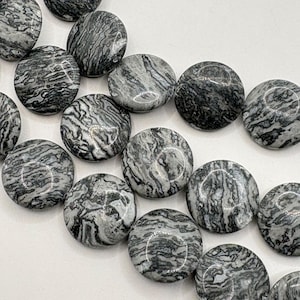 May include: Close-up of a strand of round, flat beads. The beads are a mix of gray and black with a swirled, marbled pattern. The beads are smooth and polished, reflecting light. The background is plain white.