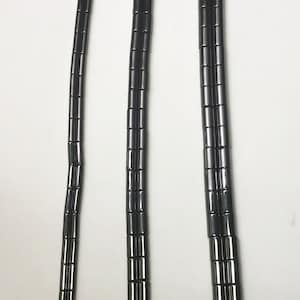 May include: Three strands of black, cylindrical beads, each strung on a red thread. The beads are evenly spaced and appear to be made of a shiny, dark material. The strands vary in length, with one having a loop at the end.