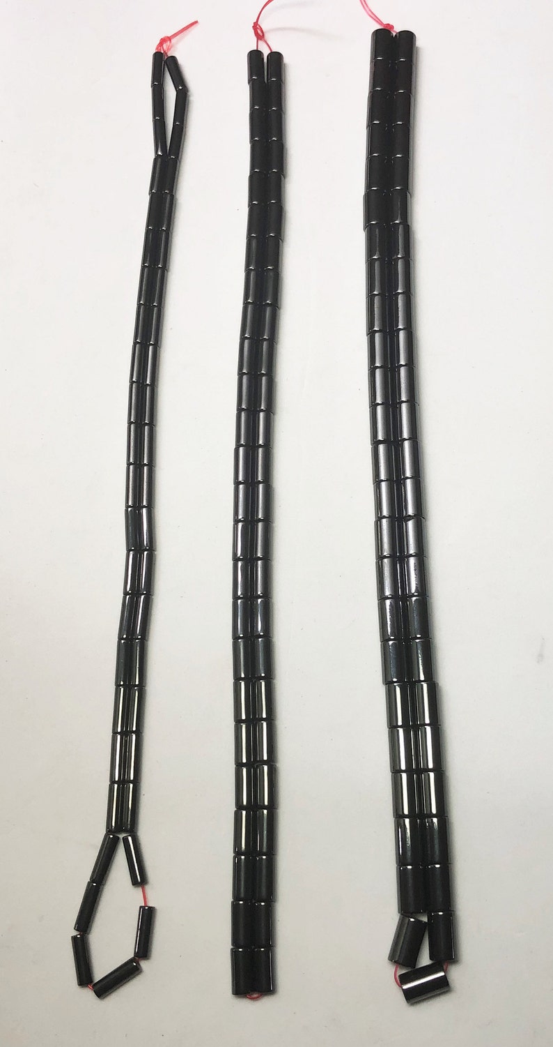 May include: Three strands of black, cylindrical beads, each strung on a red thread. The beads are evenly spaced and appear to be made of a shiny, dark material. The strands vary in length, with one having a loop at the end.