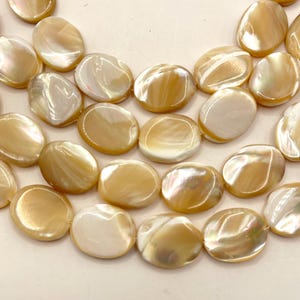 May include: Close-up of a collection of iridescent, oval-shaped beads. The beads have a creamy, golden-yellow color with shimmering, pearlescent highlights. The beads are arranged in rows, suggesting they are for jewelry making.