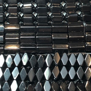 May include: Close-up of several strands of black gemstone beads. The beads are in various shapes, including faceted rectangles, cylinders, and diamond shapes. The beads have a glossy, reflective surface, and are strung together. The background is white.