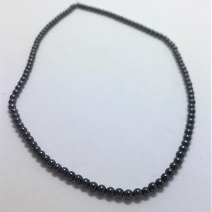 May include: A dark grey beaded necklace. The necklace is made up of small, round beads strung together to form a continuous loop. The beads have a smooth, polished appearance and reflect light, creating a subtle sheen.