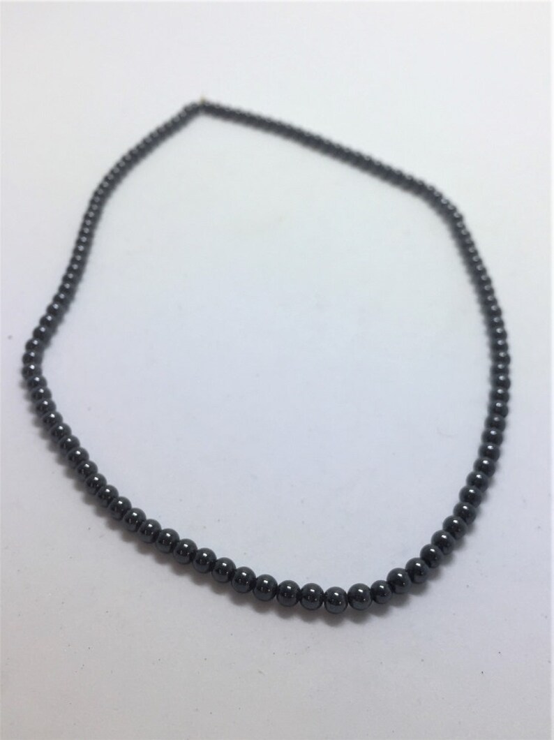May include: A dark grey beaded necklace. The necklace is made up of small, round beads strung together to form a continuous loop. The beads have a smooth, polished appearance and reflect light, creating a subtle sheen.
