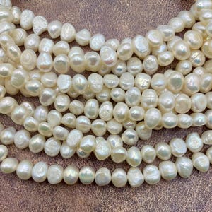 May include: Close-up of multiple strands of creamy white, irregularly shaped freshwater pearls. The pearls have a lustrous sheen and vary in size and shape, creating a natural, organic aesthetic. The background is a warm, neutral tone, highlighting the pearls' color.