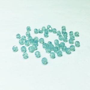 May include: A scattering of small, faceted beads in a pale blue-green hue. The beads are round with multiple cut surfaces, creating a sparkling effect. They are scattered on a white surface, suggesting a craft or jewelry-making theme.