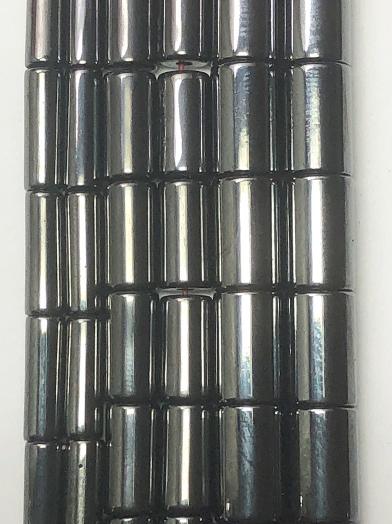 May include: Close-up of a strand of dark gray, cylindrical beads. The beads are arranged in neat rows, reflecting light and appearing smooth. The beads are likely for jewelry making.