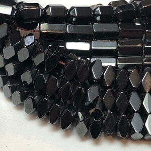 May include: Close-up of a strand of black gemstone beads. The beads are faceted in various geometric shapes, including rectangles and diamond-like forms. The beads have a high-gloss finish, reflecting light. The overall theme is jewelry and gemstones.