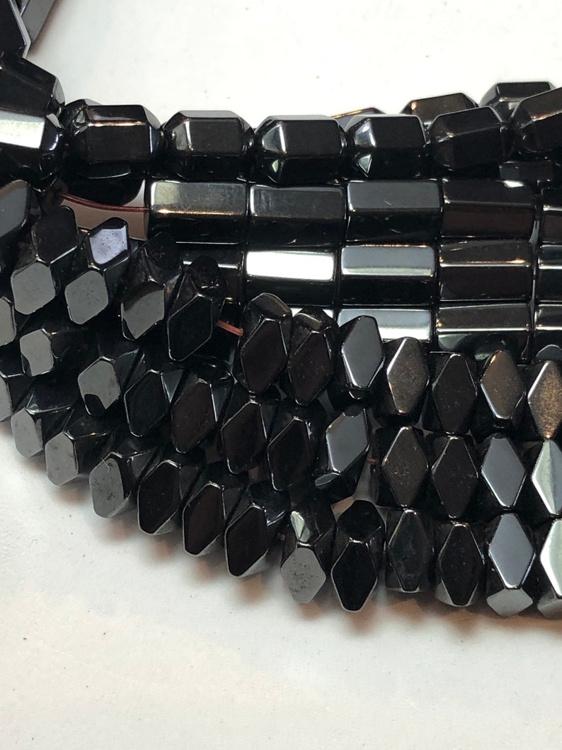 May include: Close-up of a strand of black gemstone beads. The beads are faceted in various geometric shapes, including rectangles and diamond-like forms. The beads have a high-gloss finish, reflecting light. The overall theme is jewelry and gemstones.