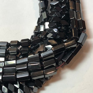 May include: Close-up of strands of black hematite beads. The beads are rectangular with faceted edges, giving them a reflective, metallic appearance. The beads are strung together, suggesting their use in jewelry making or crafting.