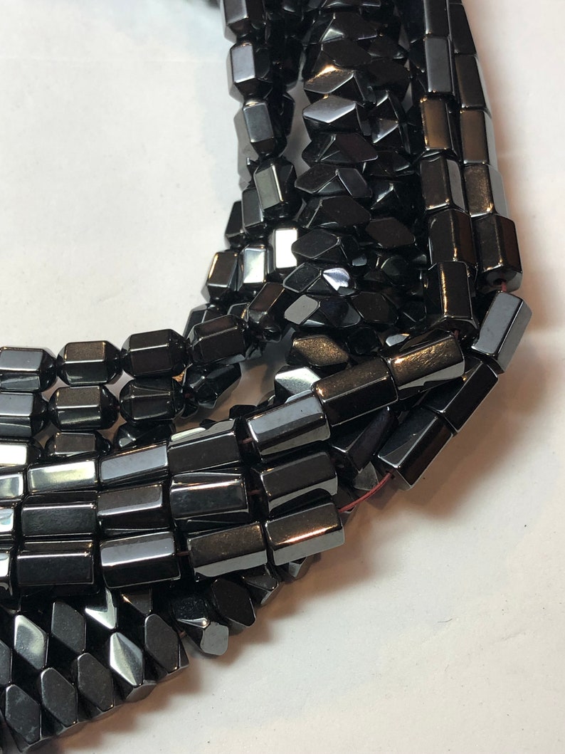May include: Close-up of strands of black hematite beads. The beads are rectangular with faceted edges, giving them a reflective, metallic appearance. The beads are strung together, suggesting their use in jewelry making or crafting.