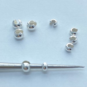 May include: A collection of small, shiny, silver-colored crimp beads and two small beads on a silver-colored tool. The crimp beads are in a variety of positions, some clustered and some in a line. The beads are used for jewelry making.