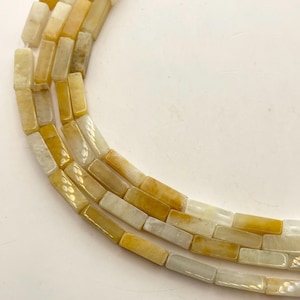 May include: Close-up of strands of rectangular beads in shades of yellow, cream, and white. The beads are strung together, creating a curved shape. The beads have a polished, smooth surface and appear to be made of stone or a similar material.