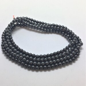May include: Multiple strands of dark grey hematite beads. The small, round beads are strung together, creating several rows. The beads have a metallic sheen and are displayed against a white background.