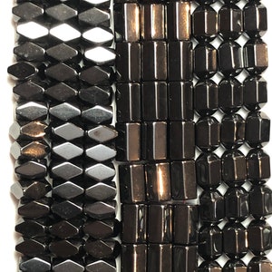 May include: Close-up of three strands of dark gray hematite beads. The beads are in various shapes: diamond, rectangular, and faceted. The beads are strung together, ready for jewelry making. The beads have a polished, reflective surface.