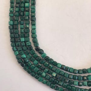 May include: Strands of dark green malachite beads. The beads are square-shaped with faceted edges, creating a textured appearance. The beads are strung together, forming multiple strands. The color varies from light to dark green.