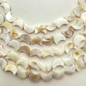May include: Close-up of a collection of crescent-shaped, iridescent beads. The beads are a mix of white, cream, and tan colors, with a pearlescent sheen. The beads are arranged in curved rows, suggesting they are for jewelry making.