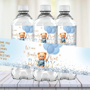 May include: Three clear plastic water bottles with blue and gold labels. The labels feature a cartoon bear holding blue balloons and the text "We can barely wait baby".