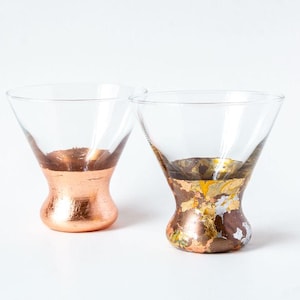 May include: Two clear martini glasses with metallic copper and gold leaf detailing on the lower half. The glasses have an inverted cone shape and are set against a white backdrop. The copper finish has a smooth, reflective surface.