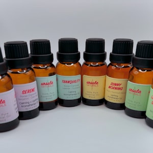 May include: Ten amber glass bottles with black caps and colorful labels. The labels have the words "SERENE", "ursula NATURALS", "TRANQUILITY", "ursula NATURALS", "SUNNY MORNING", "ursula NATURALS", "la", "S", and "A". The bottles are arranged in a semi-circle on a white surface. The bottles are filled with essential oils.