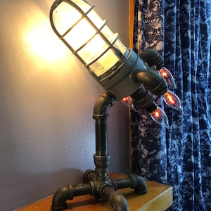 May include: A gray and black rocket-shaped lamp with a caged light bulb and a pipe base. The lamp is on a wooden shelf with a blue and white patterned curtain in the background.