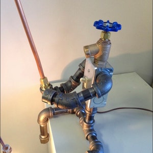 May include: A table lamp made from repurposed plumbing pipes and fittings. The lamp has a copper cord and a blue knob on the top. The lamp is sitting on a white surface.